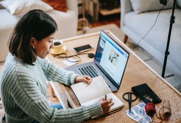 Woman with laptop in front of her taking notes in a notebook